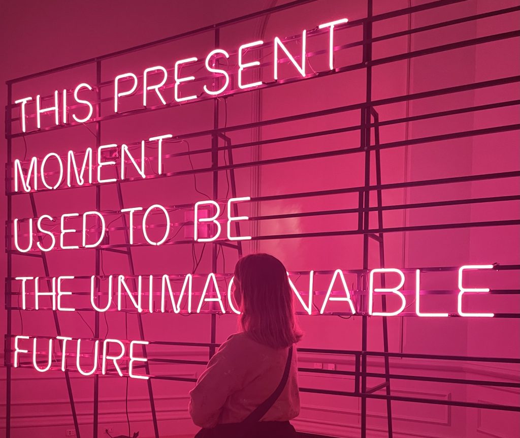 a woman standing in front of a sign that says "this present moment used to be the unimaginable future"